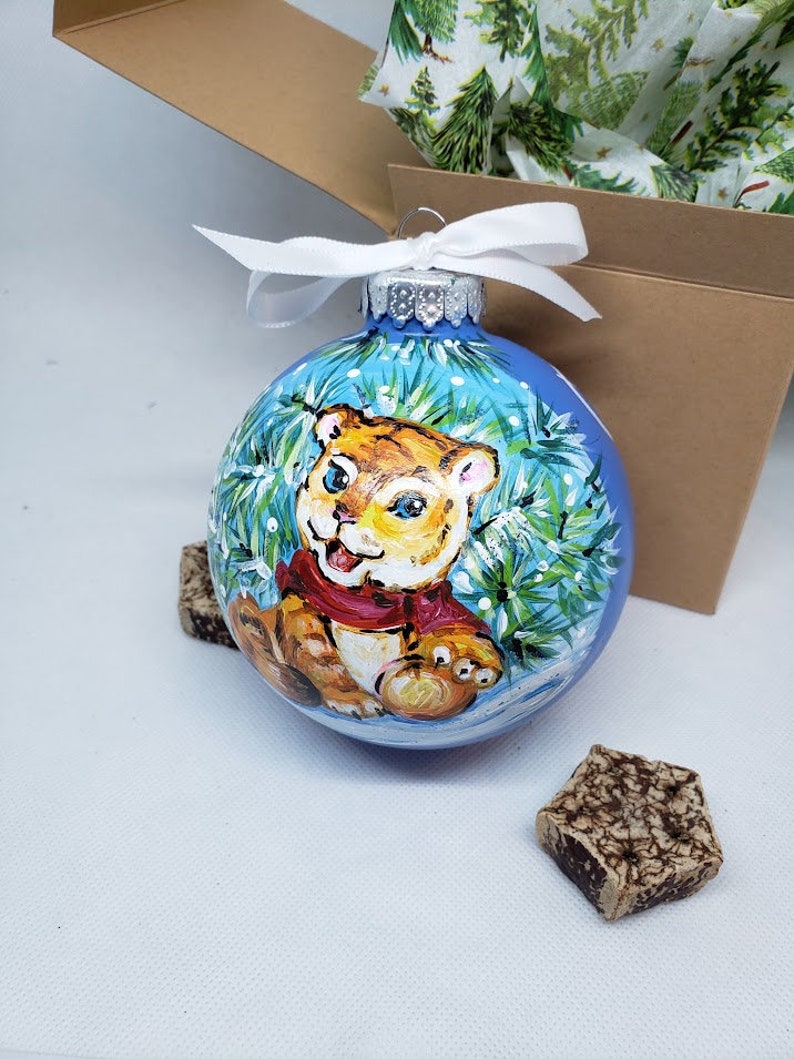 May include: A blue glass Christmas bauble with a hand-painted image of a tiger cub wearing a red scarf and surrounded by green pine branches and white snowflakes.