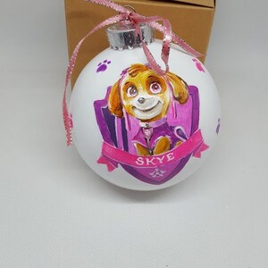 May include: A white Christmas bauble with a hand-painted image of Skye from Paw Patrol. The bauble features a purple and pink shield with paw prints and the name "Skye" written in pink.