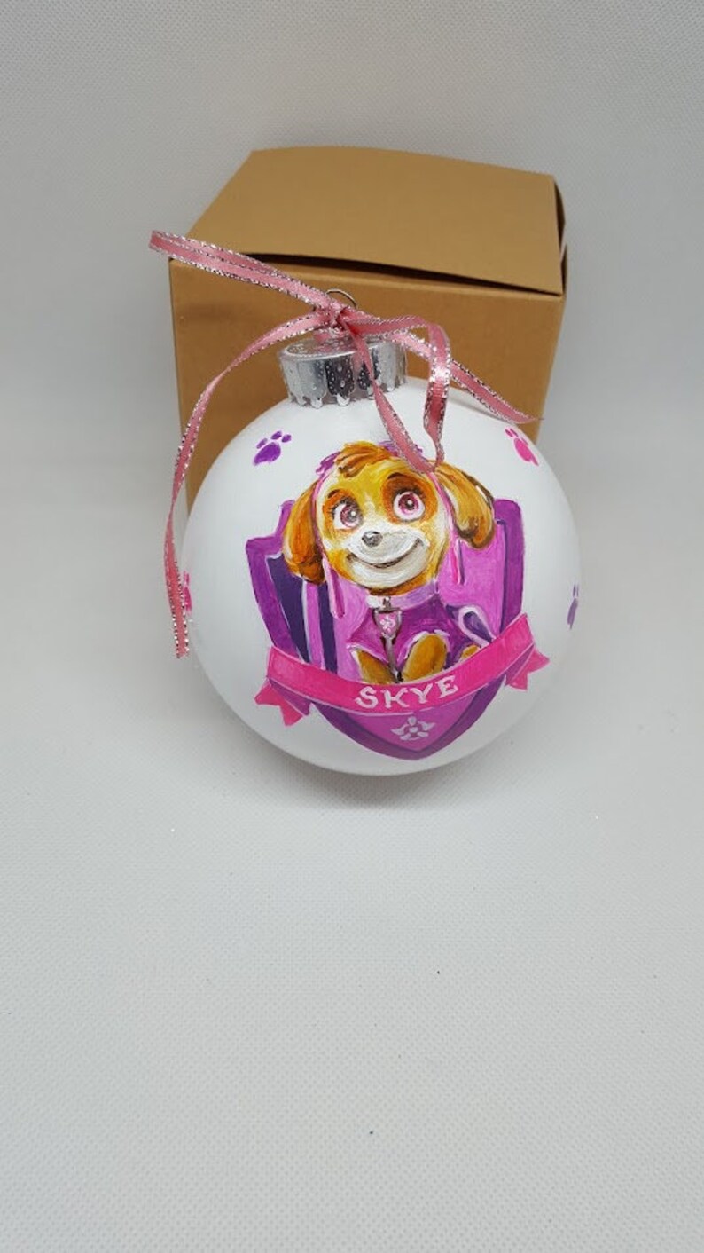 May include: A white Christmas bauble with a hand-painted image of Skye from Paw Patrol. The bauble features a purple and pink shield with paw prints and the name "Skye" written in pink.