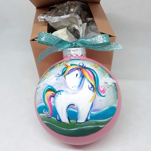 May include: A pink glass Christmas bauble featuring a hand-painted unicorn design. The unicorn is white with a rainbow mane and tail, and is standing on a green field with clouds and stars in the background.