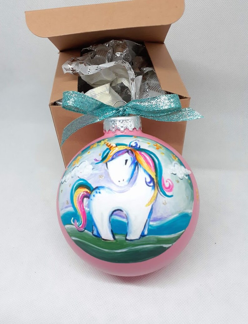 May include: A pink glass Christmas bauble featuring a hand-painted unicorn design. The unicorn is white with a rainbow mane and tail, and is standing on a green field with clouds and stars in the background.