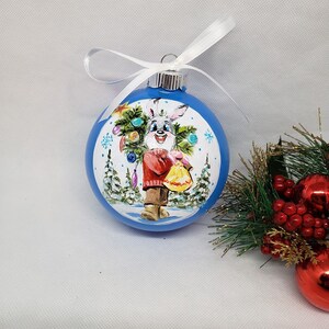 May include: A blue glass Christmas bauble with a hand-painted design of a bunny holding a small Christmas tree. The bunny is wearing a red sweater and brown trousers. The bauble is decorated with white snowflakes and a white background.