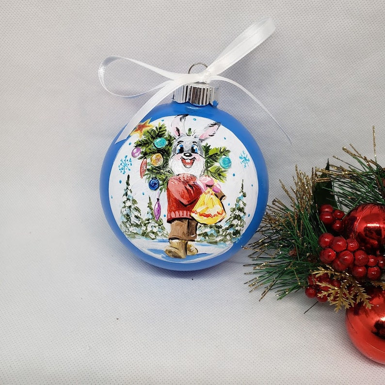 May include: A blue glass Christmas bauble with a hand-painted design of a bunny holding a small Christmas tree. The bunny is wearing a red sweater and brown trousers. The bauble is decorated with white snowflakes and a white background.