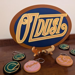 Könnte beinhalten: Ein großes, ovales Schild mit dem Text "O'DUST" in goldfarbenen Buchstaben, vor blauem Hintergrund und braunem Rand. Mehrere Schlüsselanhänger mit dem Buchstaben "C" und dem Wort "Papeles" sind ausgestellt.