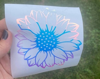 May include: A white vinyl sticker with a holographic finish depicting a sunflower with a blue center and pink and purple petals.