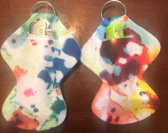 May include: Two colorful fabric keychains with a silver ring. One keychain has a silver lighter inside and the other has a green lip balm inside.
