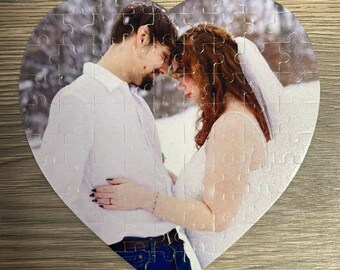 May include: A heart-shaped jigsaw puzzle featuring a couple in a snowy setting. The couple is dressed in white, with the woman wearing a veil. The puzzle is made of white pieces with a glittery finish.