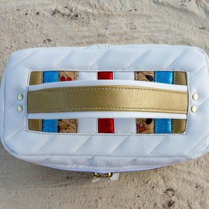 May include: White quilted leather cosmetic bag with a gold handle and colorful striped fabric accents. The bag has a zipper closure.