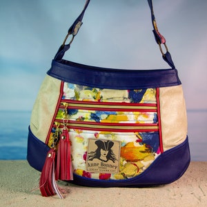 May include: A blue and beige shoulder bag with a floral print and a red tassel. The bag has a zipper closure and a label that reads "Anne Bonney, Treasure Hunter, Great Guana Cay".