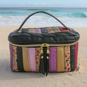 May include: A black quilted travel makeup bag with a gold zipper and tassel pulls. The bag has a striped design with pink, gold, and black stripes. The bag is sitting on a sandy beach.