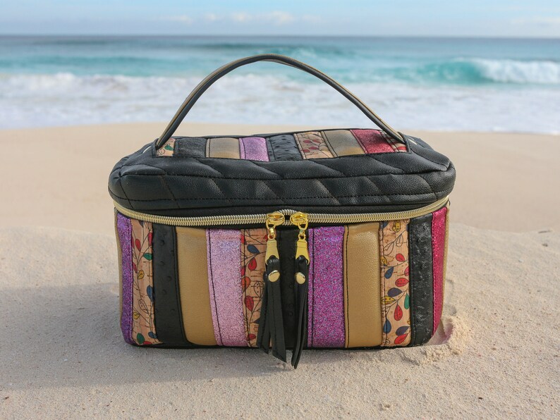 May include: A black quilted travel makeup bag with a gold zipper and tassel pulls. The bag has a striped design with pink, gold, and black stripes. The bag is sitting on a sandy beach.