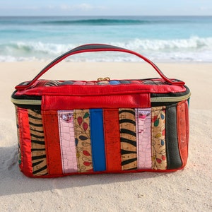 May include: A red, black, and brown striped travel bag with a zipper closure and a black handle. The bag is sitting on a white sandy beach.