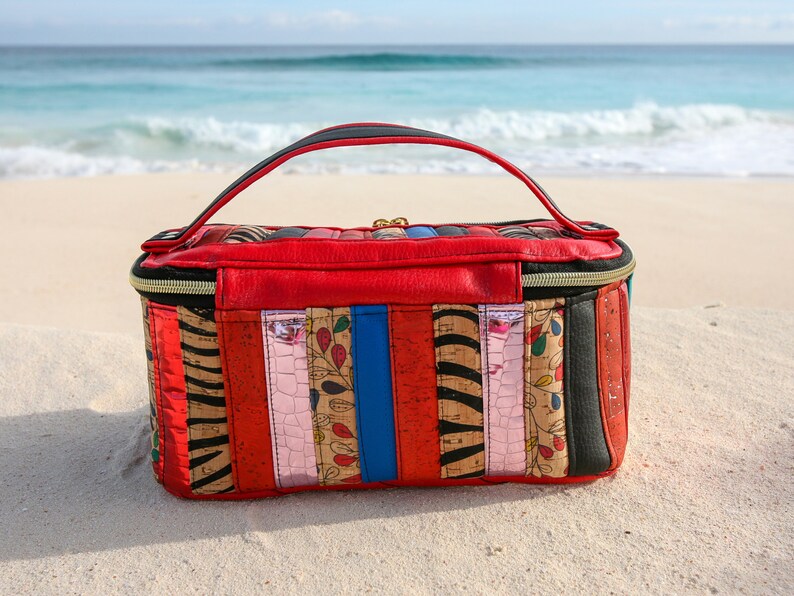 May include: A red, black, and brown striped travel bag with a zipper closure and a black handle. The bag is sitting on a white sandy beach.