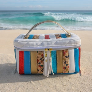 May include: A white quilted travel bag with a gold zipper and a handle. The bag has a striped design with red, blue, gold, and white stripes. The bag is sitting on a white sandy beach.
