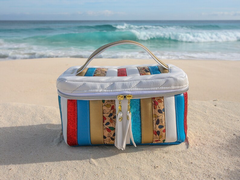 May include: A white quilted travel bag with a gold zipper and a handle. The bag has a striped design with red, blue, gold, and white stripes. The bag is sitting on a white sandy beach.