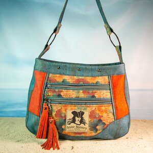 May include: A blue and orange cork fabric handbag with a floral print and a zippered pocket. The bag has a long strap and a tassel. The bag has a label that says "Anne Bonney Bahamas Great Guana Cay".