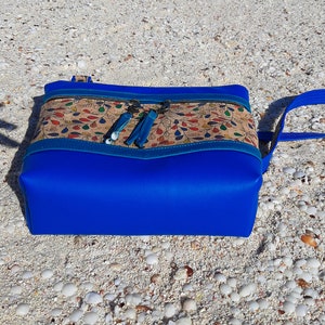 May include: A blue and brown crossbody bag with a cork fabric design and blue tassels. The bag is sitting on a white sandy beach.