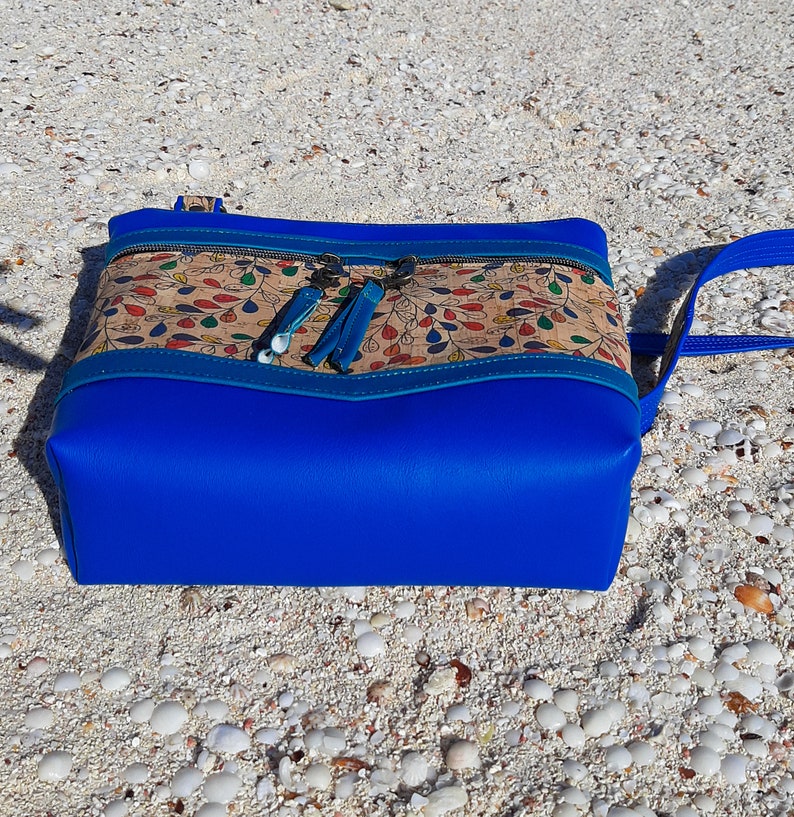 May include: A blue and brown crossbody bag with a cork fabric design and blue tassels. The bag is sitting on a white sandy beach.