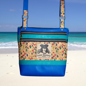 May include: A blue crossbody bag with a cork fabric front featuring a colorful leaf pattern and a patch with a pirate illustration and the text "Anne Bonney Bahamas Great Guana Cay".