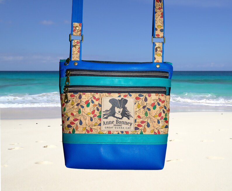 May include: A blue crossbody bag with a cork fabric front featuring a colorful leaf pattern and a patch with a pirate illustration and the text "Anne Bonney Bahamas Great Guana Cay".