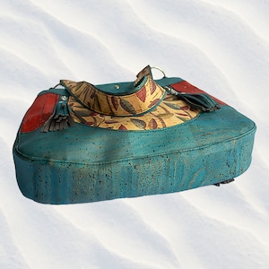 May include: A teal blue cork handbag with a yellow and red floral pattern. The bag has a long strap and a tassel on the side.