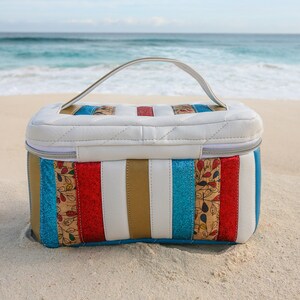 May include: A white quilted travel bag with a top handle. The bag has a striped design with red, blue, gold, and white stripes. The bag is sitting on a sandy beach.