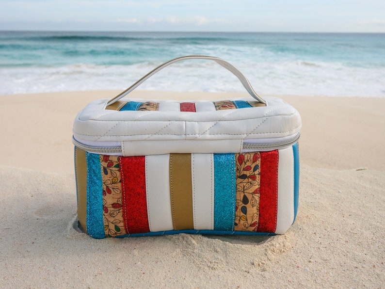 May include: A white quilted travel bag with a top handle. The bag has a striped design with red, blue, gold, and white stripes. The bag is sitting on a sandy beach.