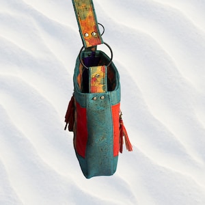 May include: A turquoise and red cork handbag with a colorful patterned strap and tassels. The bag has a top handle and a shoulder strap.