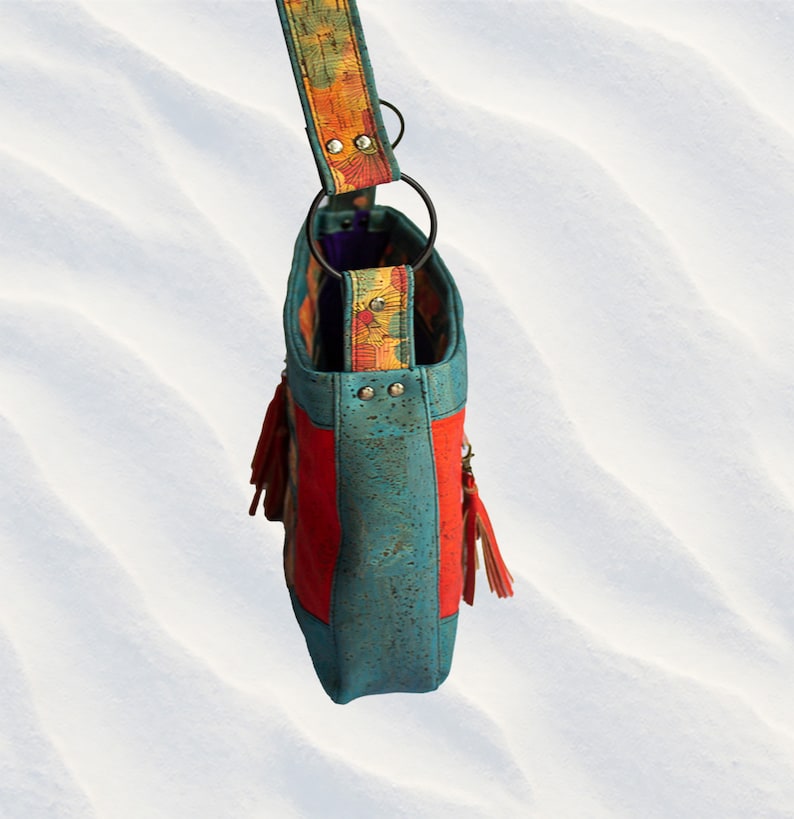 May include: A turquoise and red cork handbag with a colorful patterned strap and tassels. The bag has a top handle and a shoulder strap.