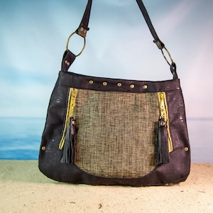 May include: A black and brown cork fabric shoulder bag with gold zipper accents and tassels. The bag has a woven fabric panel in the center.