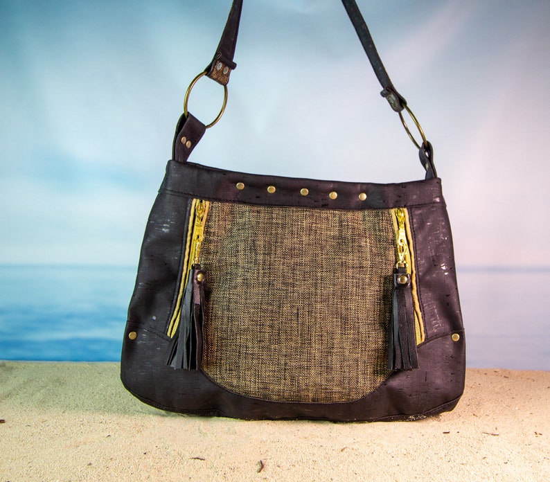 May include: A black and brown cork fabric shoulder bag with gold zipper accents and tassels. The bag has a woven fabric panel in the center.