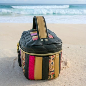 May include: A black quilted zippered travel bag with a gold zipper and a colorful striped pattern. The bag has a top handle and is sitting on a sandy beach.
