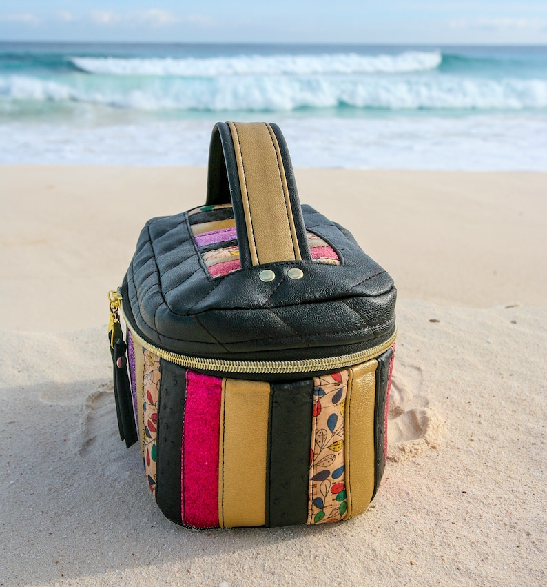 May include: A black quilted zippered travel bag with a gold zipper and a colorful striped pattern. The bag has a top handle and is sitting on a sandy beach.