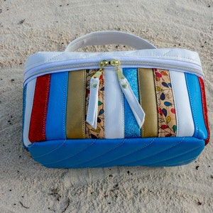 May include: A white and blue striped cosmetic bag with a zipper closure. The bag has a quilted design and is made of leather. The bag is sitting on a sandy surface.