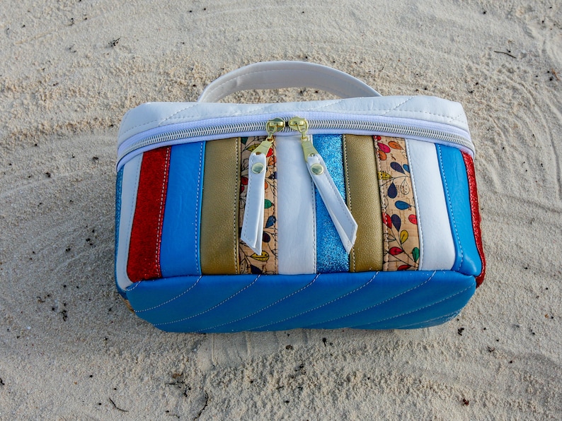 May include: A white and blue striped cosmetic bag with a zipper closure. The bag has a quilted design and is made of leather. The bag is sitting on a sandy surface.