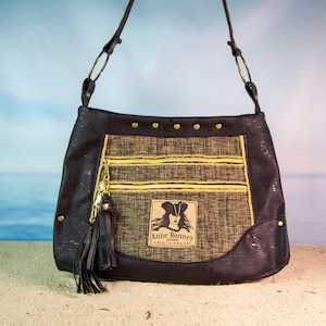 May include: A black cork handbag with a gold zipper and a gold tassel. The bag has a gold patch with the text "Anne Bonney Pirate" and "Great Guana Cay".