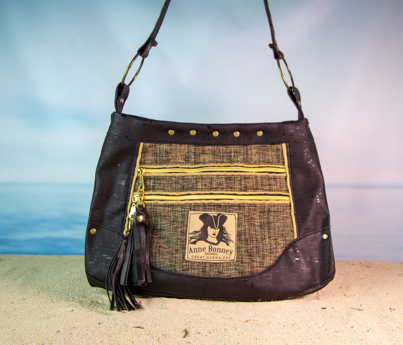 May include: A black cork handbag with a gold zipper and a gold tassel. The bag has a gold patch with the text "Anne Bonney Pirate" and "Great Guana Cay".