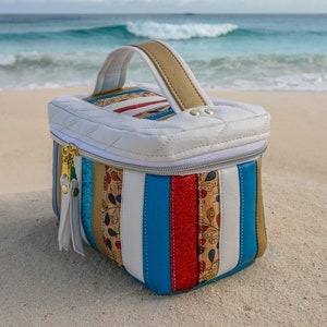 May include: A white quilted travel case with a gold handle and a zipper closure. The case is decorated with colorful stripes and a floral pattern.