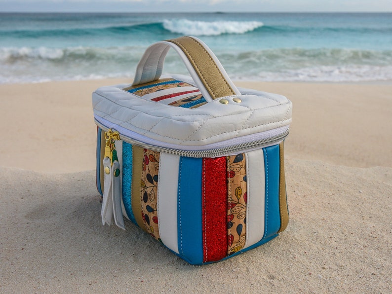 May include: A white quilted travel case with a gold handle and a zipper closure. The case is decorated with colorful stripes and a floral pattern.