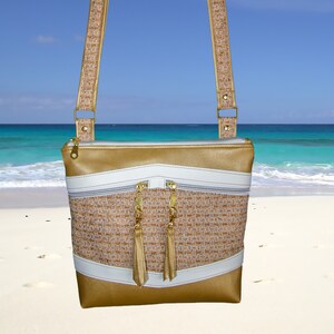 May include: A gold and white crossbody bag with a woven fabric design and gold tassel zipper pulls. The bag is photographed against a beach background.