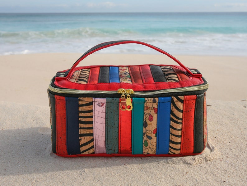 May include: A red and black striped travel makeup bag with a gold zipper and a black handle. The bag is sitting on a sandy beach.