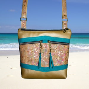 May include: A gold and turquoise crossbody bag with a cork-like texture and two zippered pockets. The bag has a long strap and is photographed against a beach background.