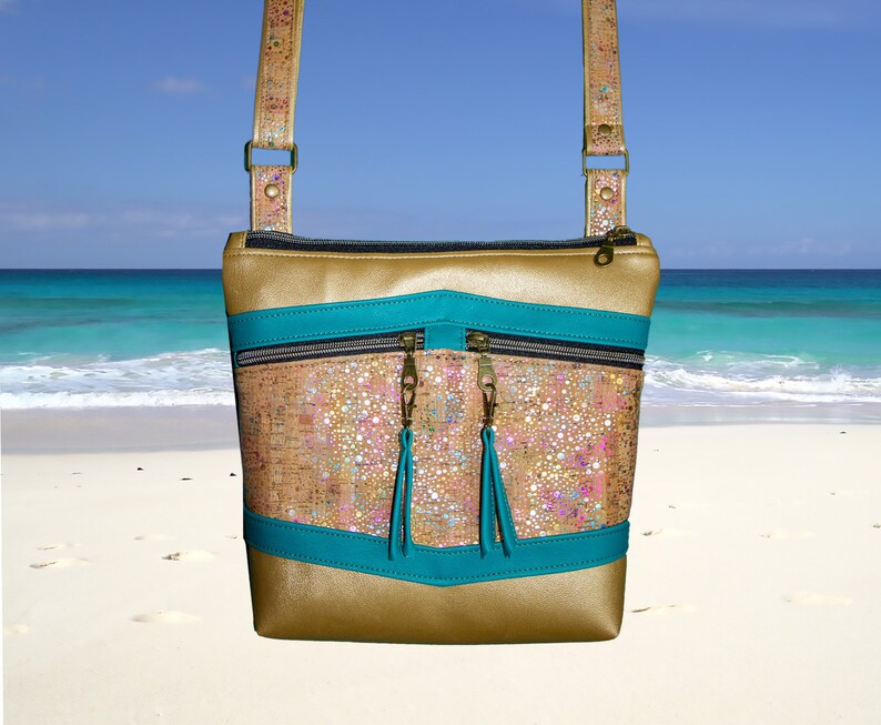 May include: A gold and turquoise crossbody bag with a cork-like texture and two zippered pockets. The bag has a long strap and is photographed against a beach background.