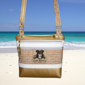 May include: A gold and white crossbody bag with a woven texture and a zipper closure. The bag features a patch with a pirate woman illustration and the text "Anne Bonney, Bahamas, Great Guana Cay".