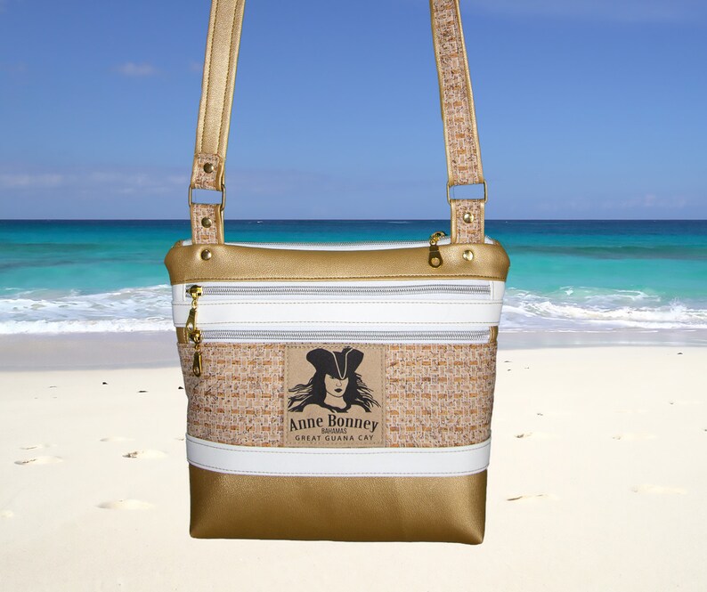 May include: A gold and white crossbody bag with a woven texture and a zipper closure. The bag features a patch with a pirate woman illustration and the text "Anne Bonney, Bahamas, Great Guana Cay".