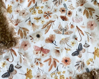 Puede incluir: Una suave manta blanca para bebé con estampado floral y mariposas. La manta tiene un respaldo de piel sintética marrón.