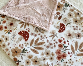 Puede incluir: Una manta suave y mullida con fondo blanco y un patrón floral en tonos marrones, rosas y rojos. La manta presenta pequeñas flores, hojas y setas. El reverso de la manta es de color rosa sólido.