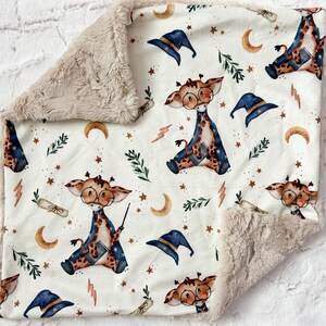 May include: A white baby blanket with a soft, plush texture on one side and a printed cotton fabric on the other. The printed side features a whimsical pattern of giraffes wearing wizard hats and holding wands, surrounded by stars, moons, and leaves.