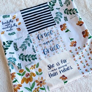 May include: A white fleece blanket with a floral pattern and the text "Grace upon Grace" and "She is far more precious than rubies." The blanket is made of soft, comfortable fabric and is ideal for snuggling up on the sofa.