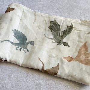 May include: A white muslin swaddle blanket with a repeating pattern of dragons and crescent moons. The dragons are in shades of green, brown, and beige.